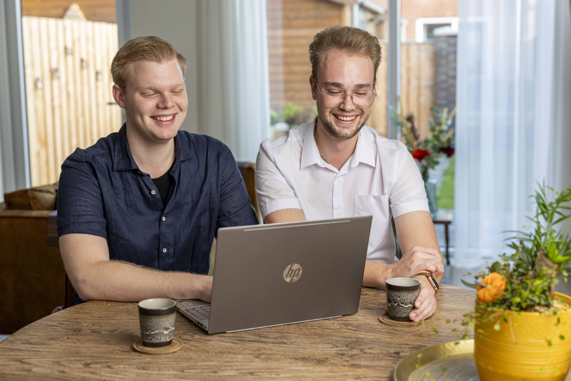 Twee lachende mannen aan tafel met een laptop en koffiekopjes.